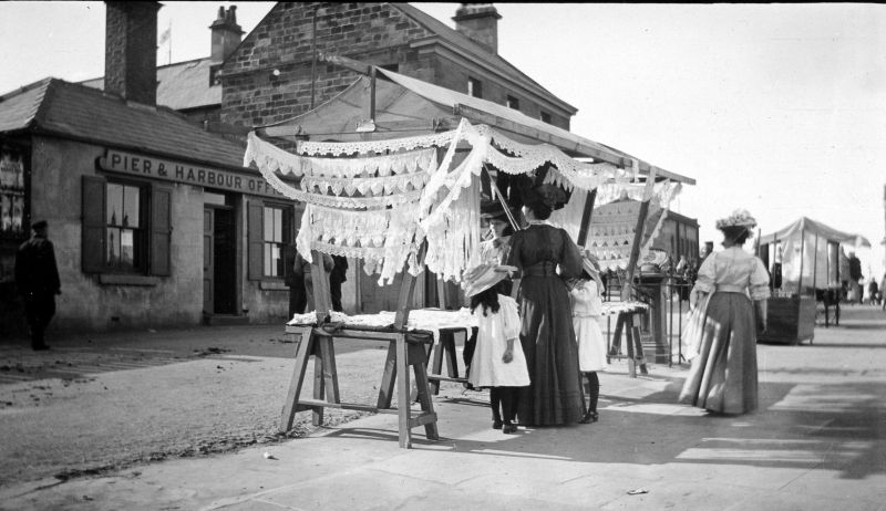 This photograph taken along Pier Road in the early 20th century however, is evidence that by now it must have been possible to set up stalls and sell goods elsewhere in the town than in the Market Place. A display of different breadths of lace has attracted the attention of a well-dressed lady with two small daughters at her side. It seems that this lace stall operated on this spot during the summer months during the Edwardian period and up to the outbreak of the First World War. (Courtesy of: Whitby Museum, Photographic Collection, W 4394. Unknown photographer).